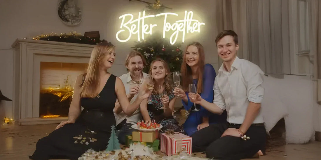 Group of five friends sitting by Christmas tree with gifts and a fireplace, holding wine glasses under neon Better Together sign