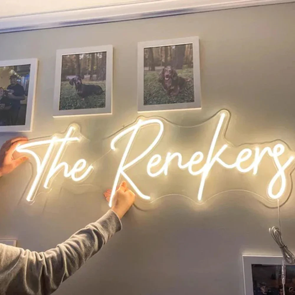 Hands mounting a glowing white neon sign that reads The Renekers on a gray wall with framed photos